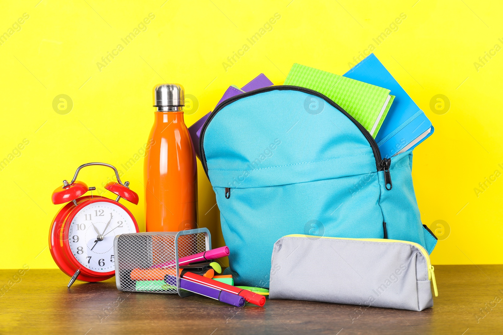 Photo of Backpack with different school stationery, bottle and alarm clock on wooden table against yellow background