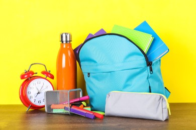 Backpack with different school stationery, bottle and alarm clock on wooden table against yellow background