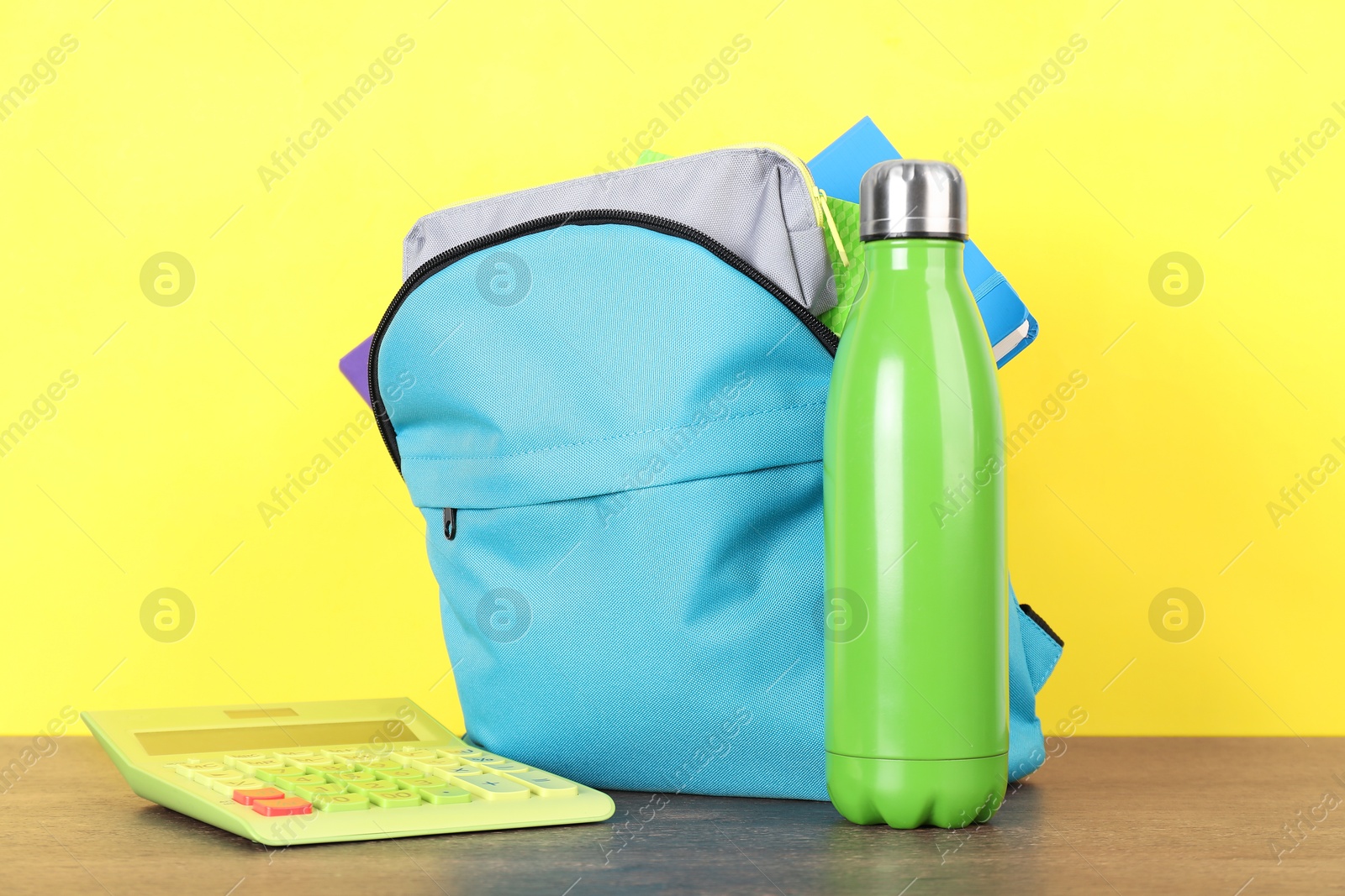 Photo of Backpack with different school stationery and bottle on wooden table against yellow background