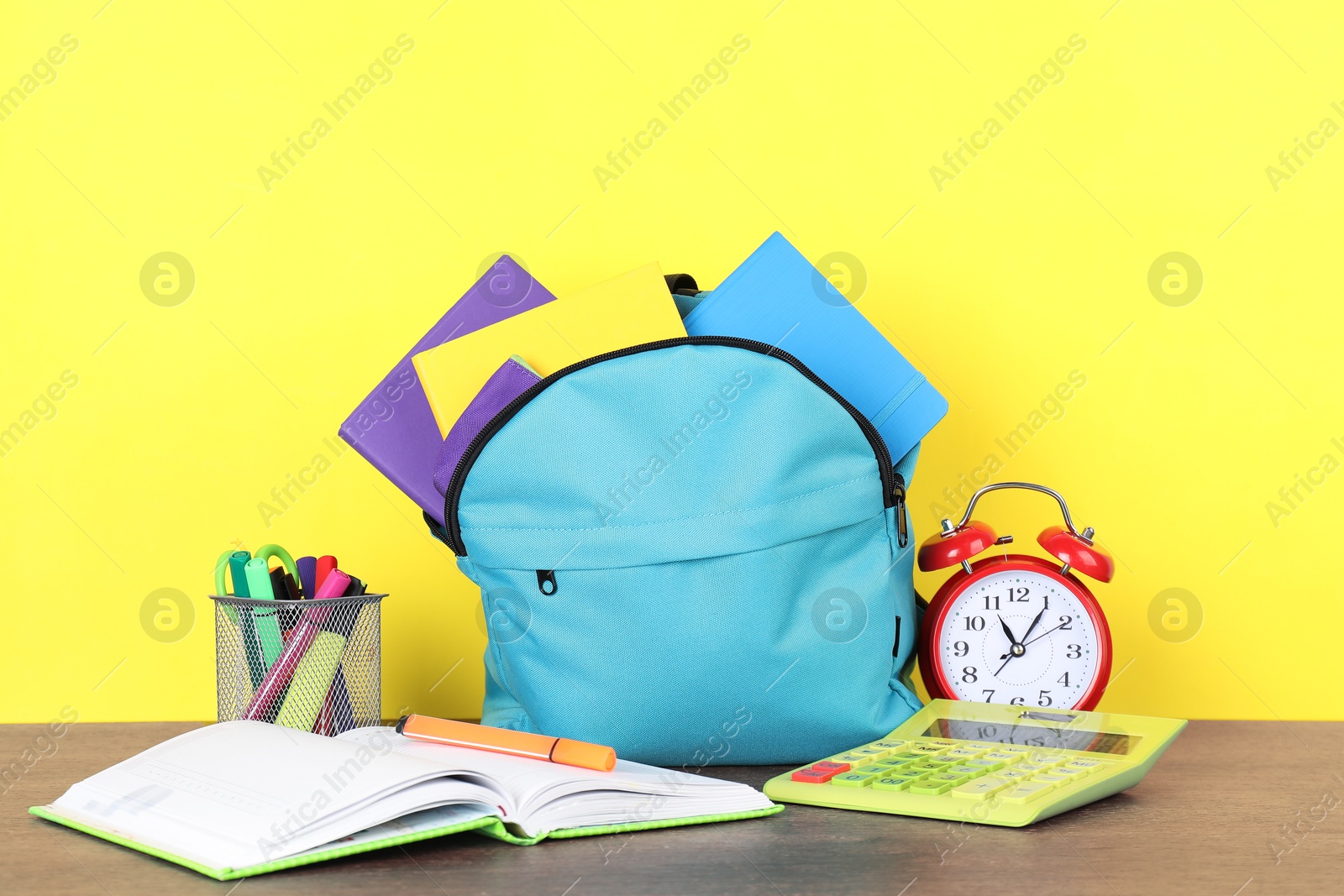 Photo of Backpack with different school stationery and alarm clock on wooden table against yellow background