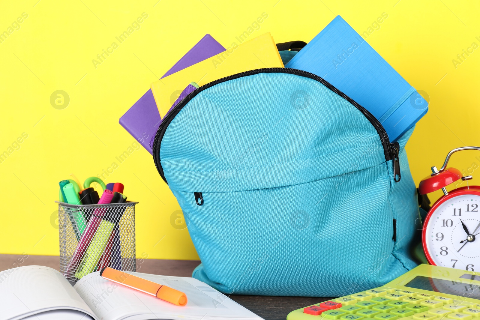 Photo of Backpack with different school stationery and alarm clock on wooden table against yellow background