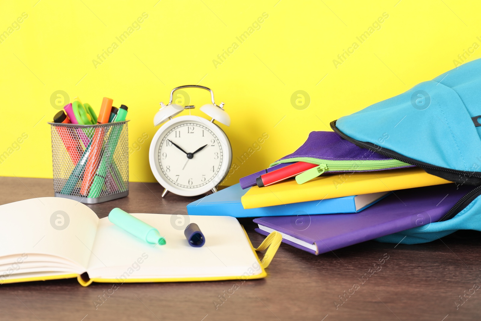 Photo of Backpack with different school stationery and alarm clock on wooden table against yellow background