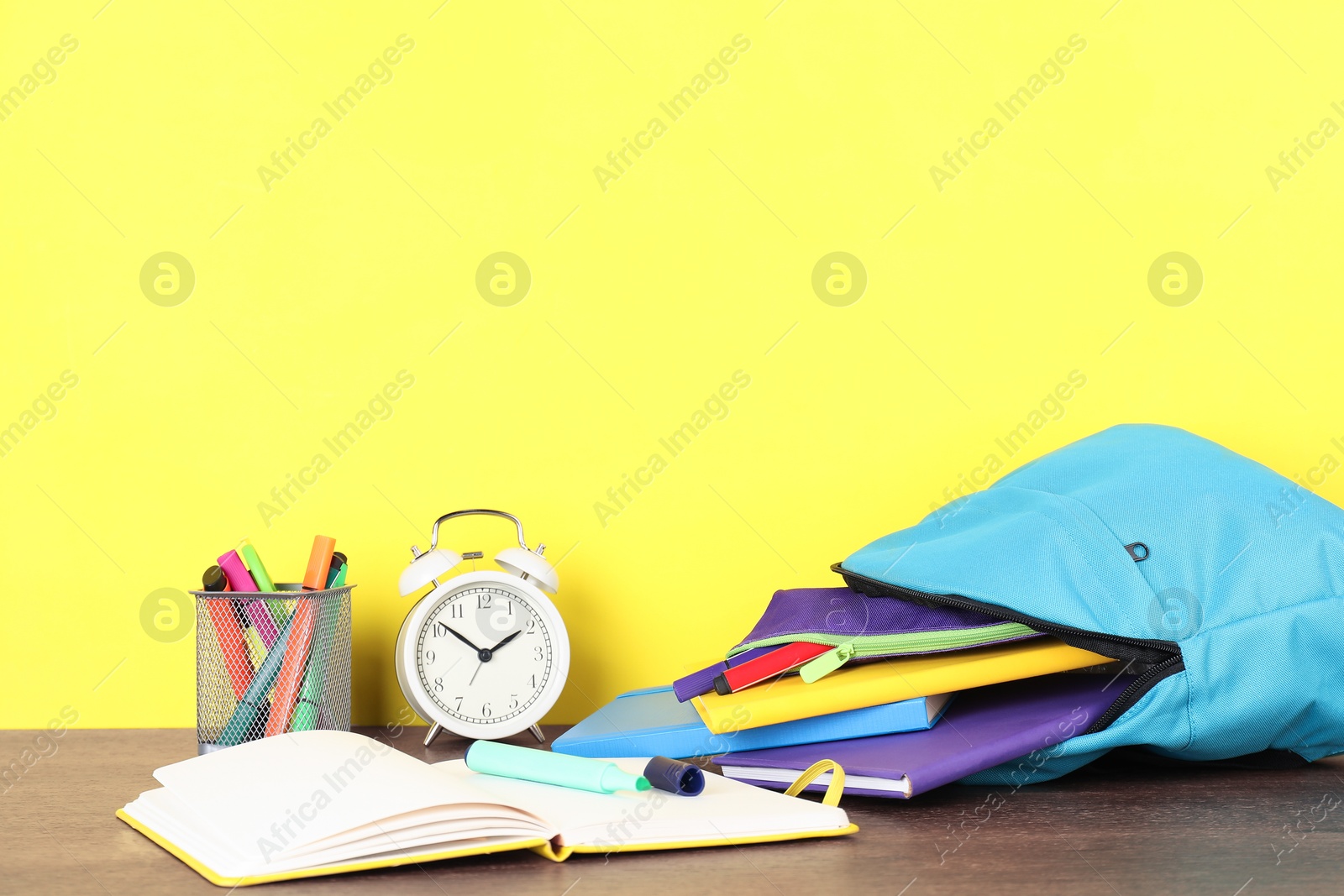 Photo of Backpack with different school stationery and alarm clock on wooden table against yellow background