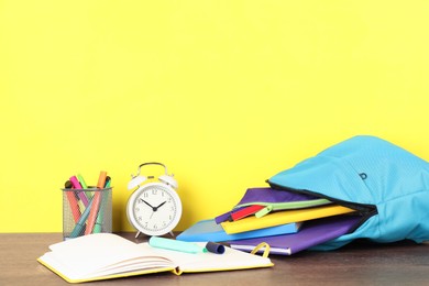 Photo of Backpack with different school stationery and alarm clock on wooden table against yellow background