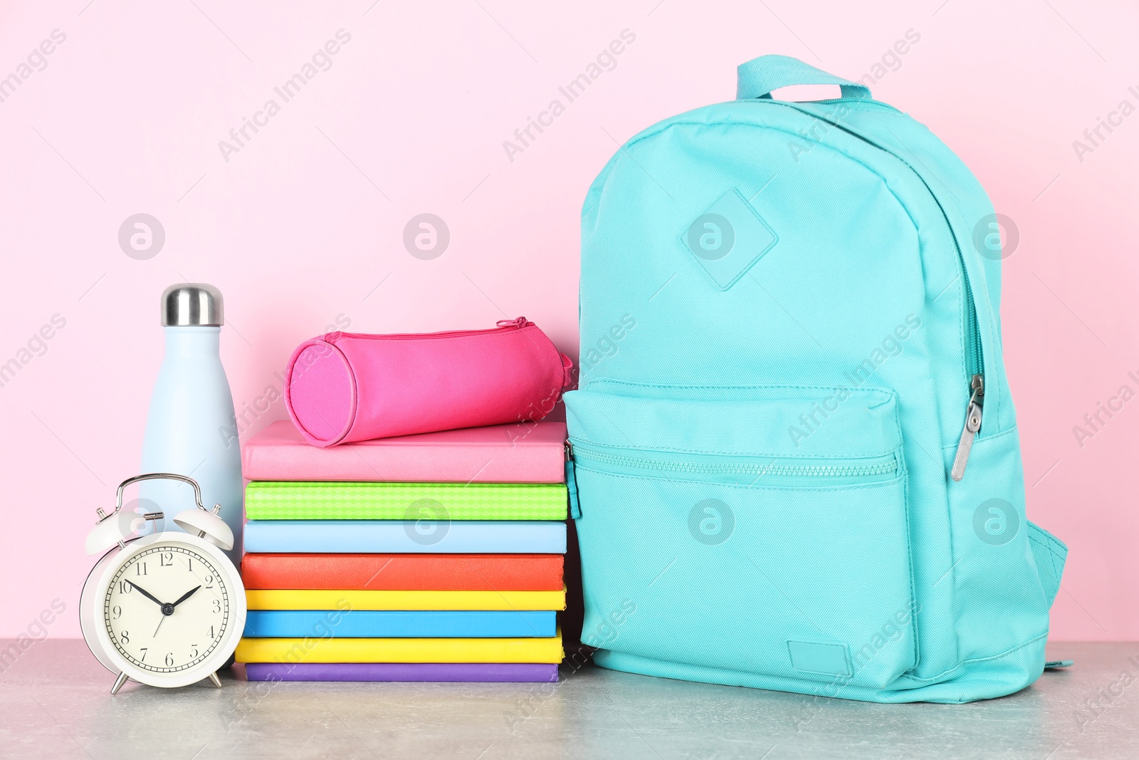 Photo of Backpack with different school stationery, alarm clock and bottle on light grey table against pink background