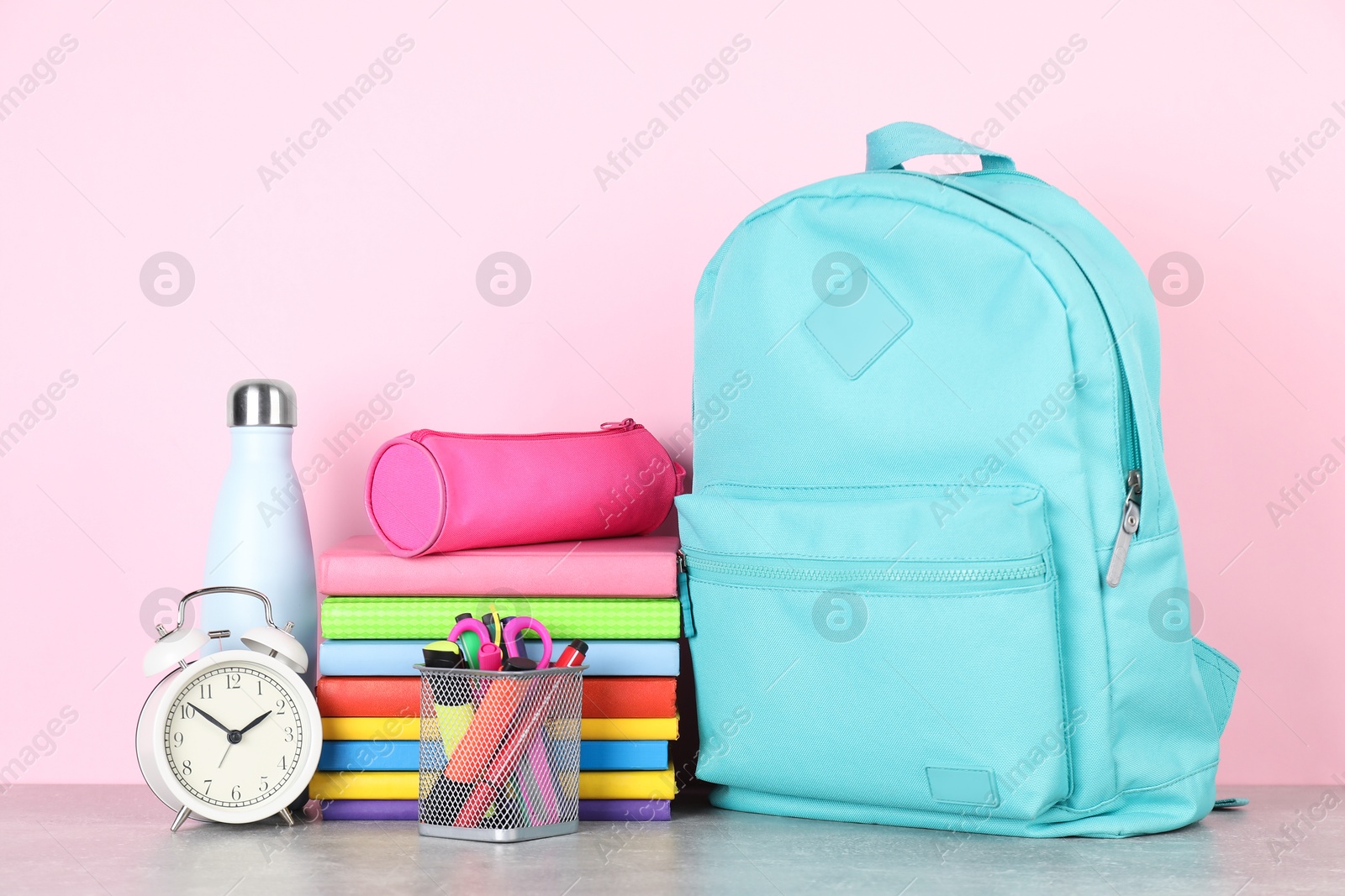 Photo of Backpack with different school stationery, alarm clock and bottle on light grey table against pink background