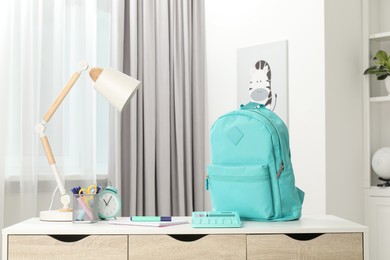 Photo of Backpack with different school stationery and alarm clock on desk in room