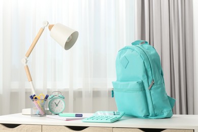 Photo of Backpack with different school stationery and alarm clock on desk in room