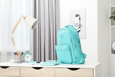 Photo of Backpack with different school stationery and alarm clock on desk in room