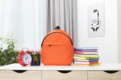 Photo of Backpack with different school stationery and alarm clock on desk in room