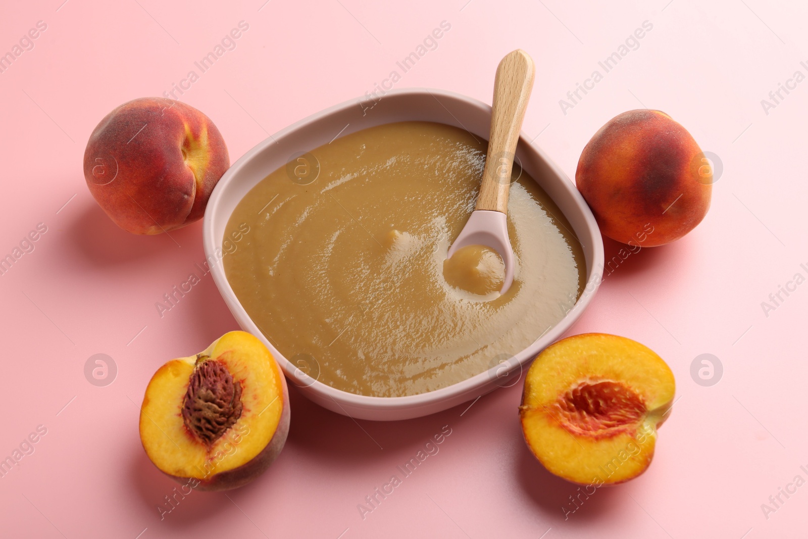 Photo of Delicious baby food with spoon in bowl and fresh peaches on pink table