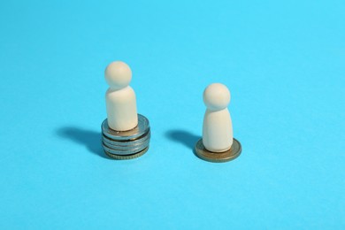 Financial inequality. One wooden figure on bigger stack of coins than another on light blue background