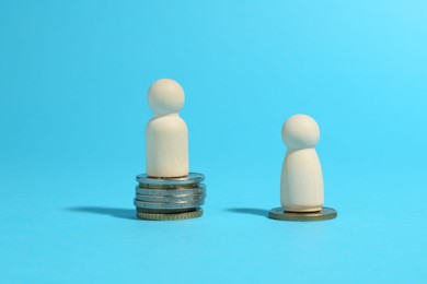 Financial inequality. One wooden figure on bigger stack of coins than another on light blue background