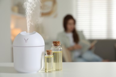 Image of Aroma diffuser and bottles of essential oil on table, space for text. Woman reading book in room, selective focus