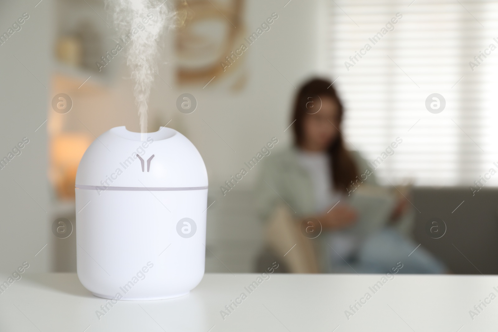 Image of Essential oil diffuser on table, space for text. Woman reading book in room, selective focus