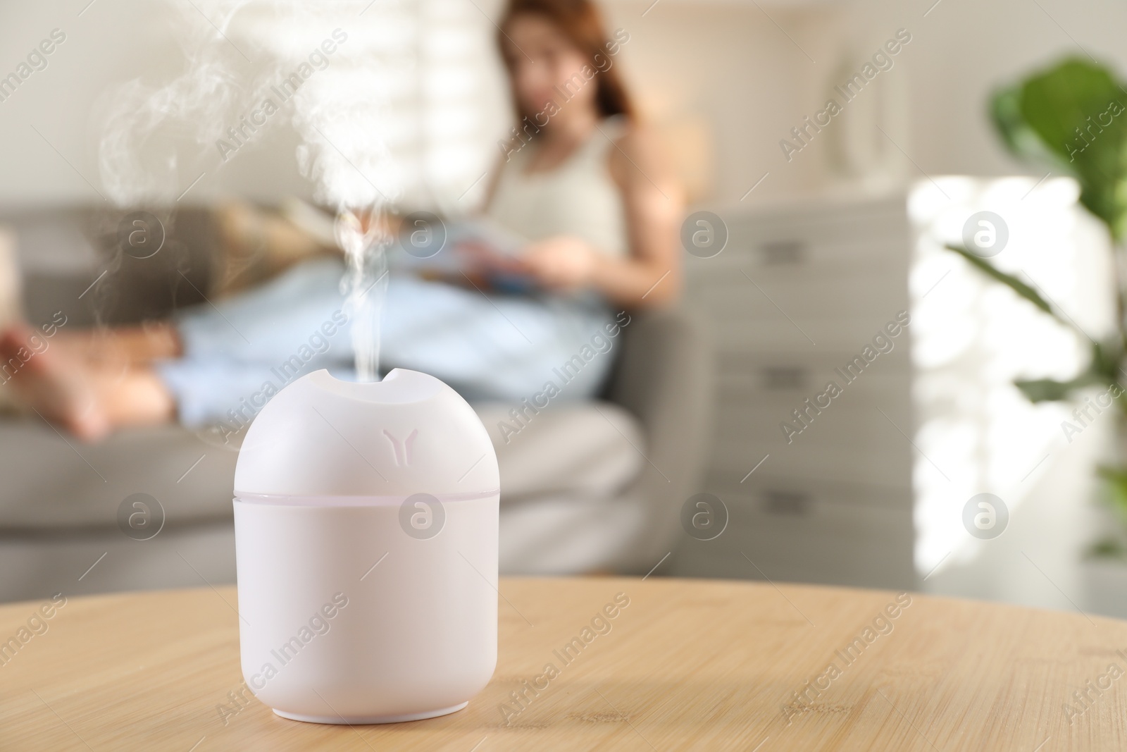 Image of Essential oil diffuser on table, space for text. Woman reading book in room, selective focus