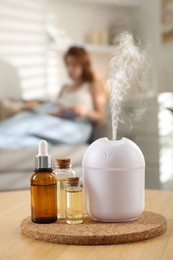 Aroma diffuser and bottles of essential oils on table, space for text. Woman reading book in room, selective focus