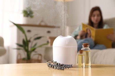 Aroma diffuser, bottles of essential oil and lavender flowers on table, space for text. Woman reading book in room, selective focus