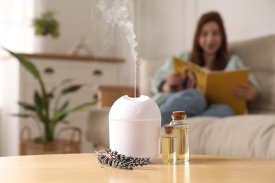 Aroma diffuser, bottles of essential oil and lavender flowers on table. Woman reading book in room, selective focus