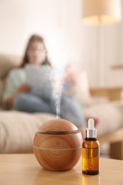 Aroma diffuser and bottle of essential oil on table, space for text. Woman reading book in room, selective focus