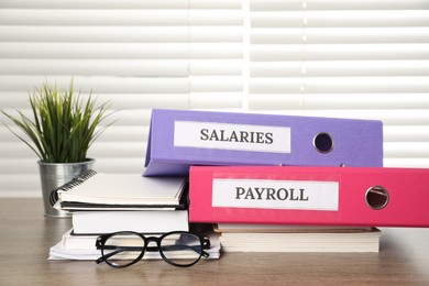 Photo of Office folders with words Payroll and Salaries, houseplant, glasses and stationery on wooden table