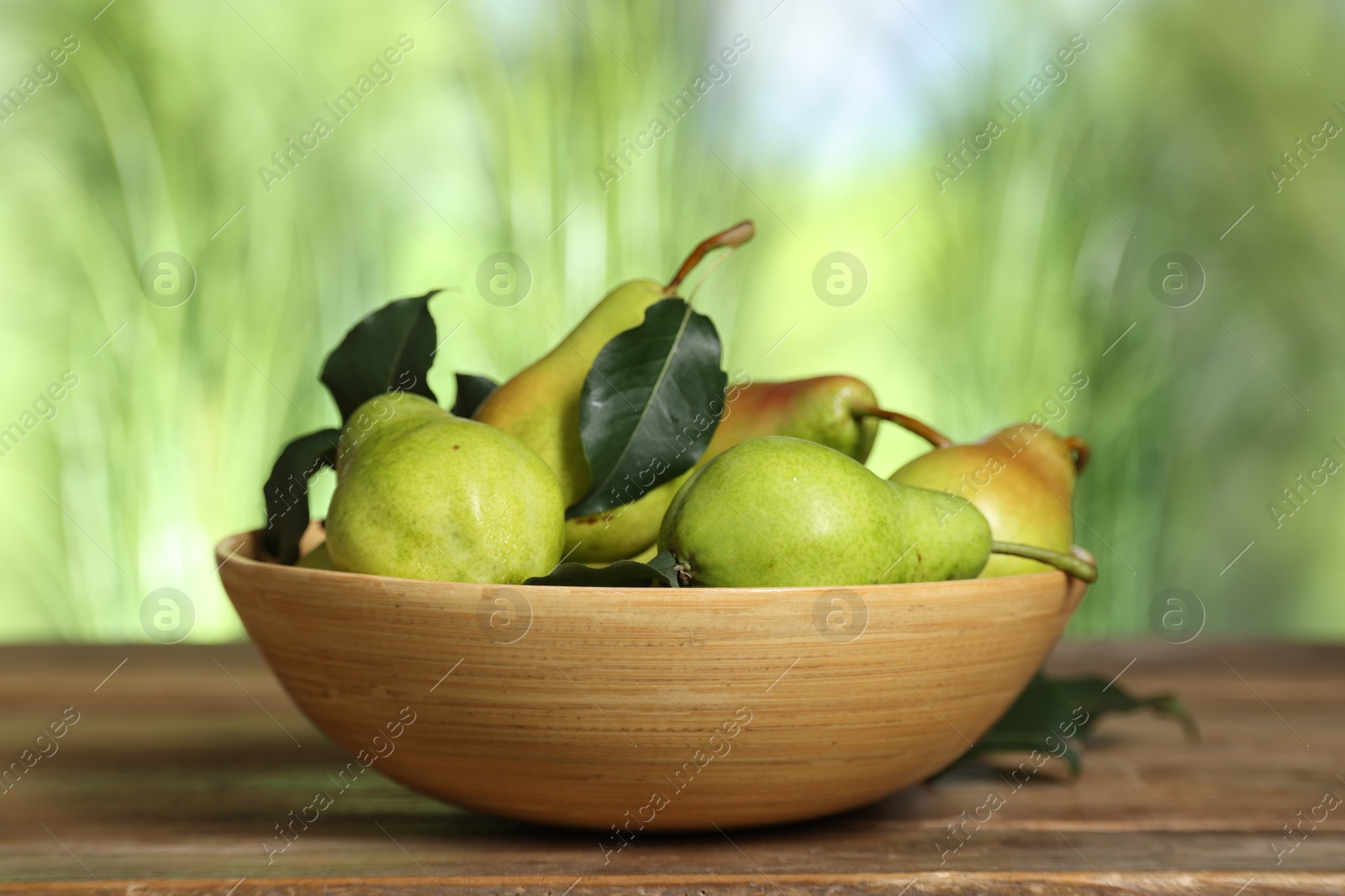 Photo of Fresh pears and leaves in bowl on wooden table against blurred green background, closeup