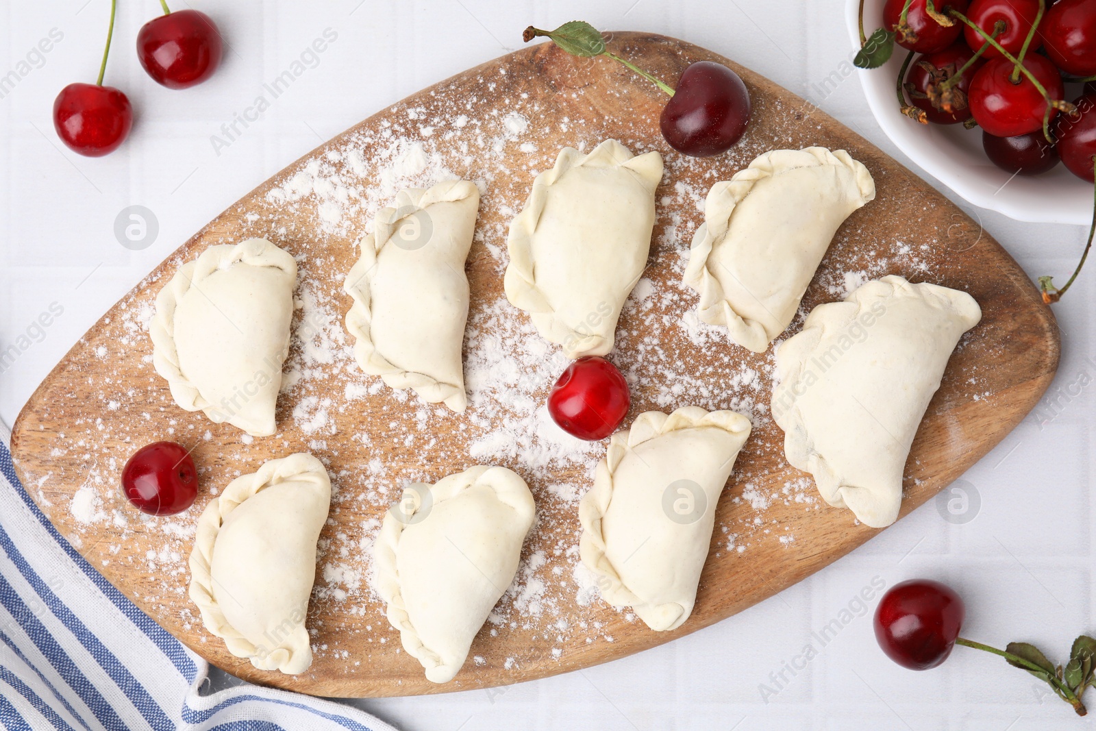 Photo of Raw dumplings (varenyky) and fresh cherries on white tiled table, flat lay