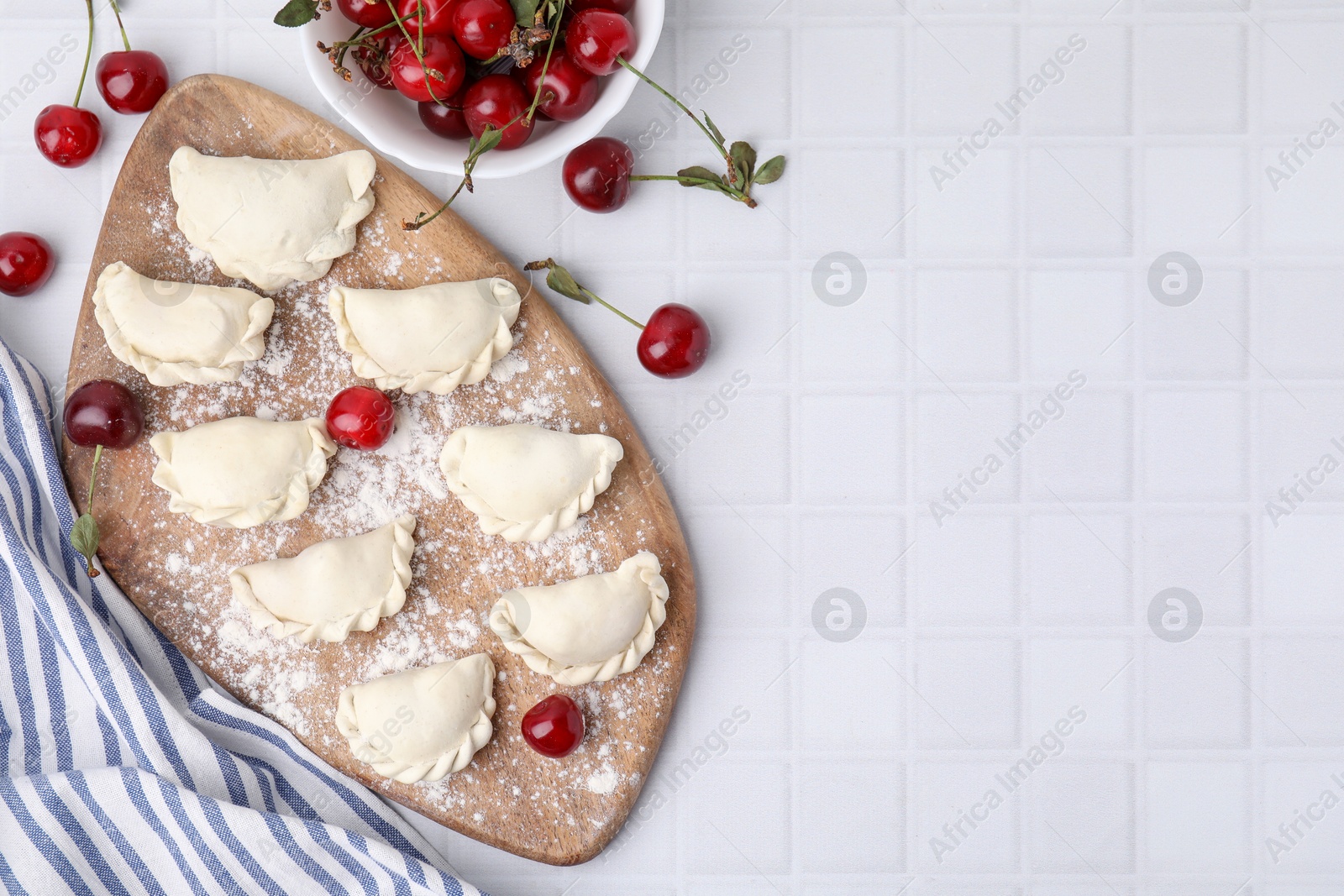 Photo of Raw dumplings (varenyky) and fresh cherries on white tiled table, flat lay. Space for text