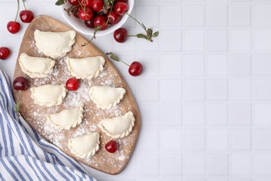 Photo of Raw dumplings (varenyky) and fresh cherries on white tiled table, flat lay. Space for text