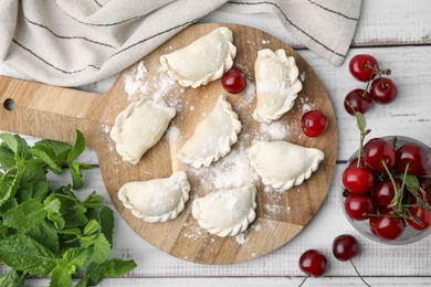 Photo of Raw dumplings (varenyky) and fresh cherries on white wooden table, flat lay