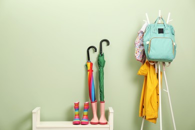 Photo of Colorful umbrellas, rubber boots and rack with jackets, backpack near green wall, space for text