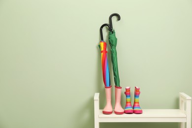 Colorful umbrellas and rubber boots on bench near green wall, space for text