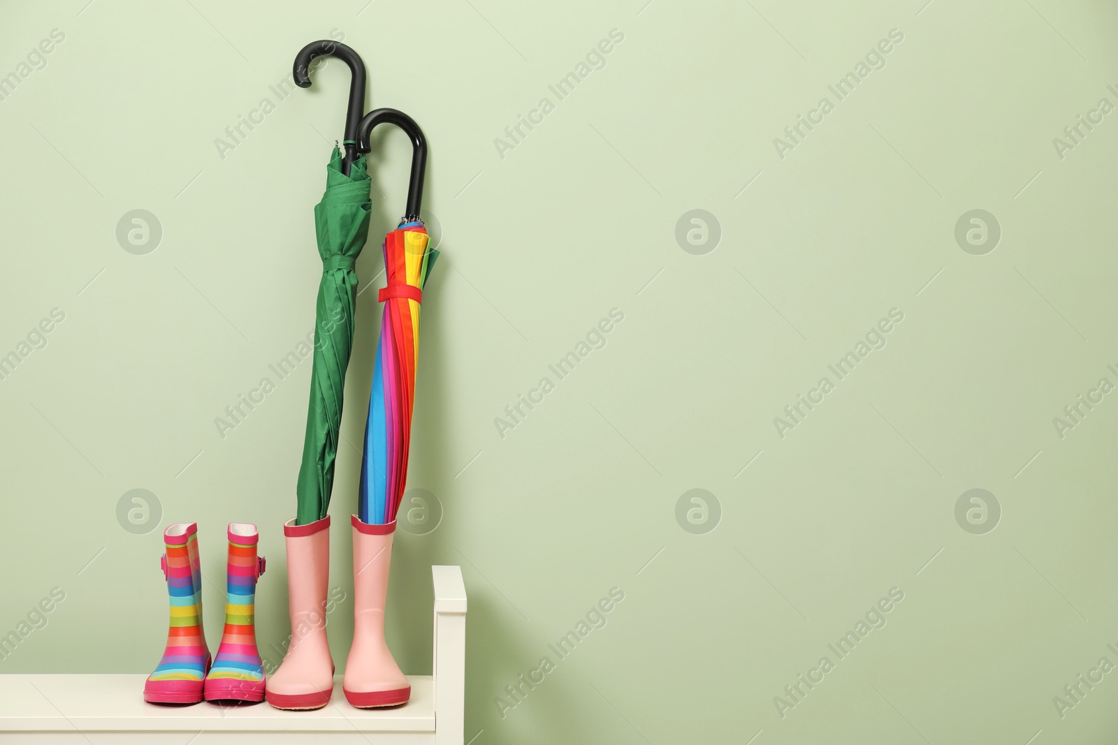 Photo of Colorful umbrellas and rubber boots on bench near green wall, space for text