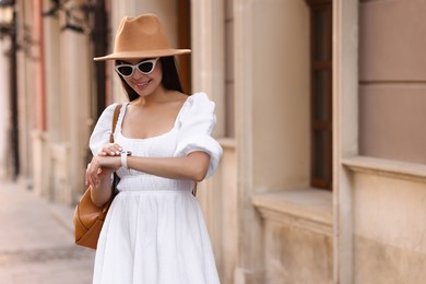 Smiling woman in stylish hat and sunglasses checking time on city street, space for text