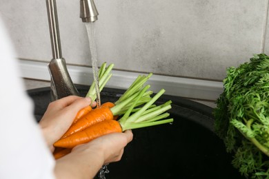 Photo of Woman washing fresh carrots under tap water in above sink indoors, closeup