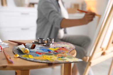 Photo of Woman painting on easel indoors, focus on palette and brushes