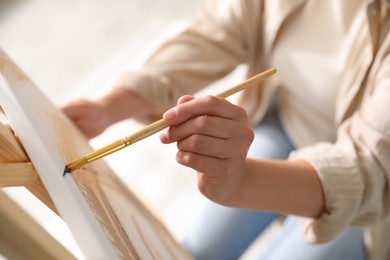 Photo of Woman painting on easel with canvas indoors, closeup