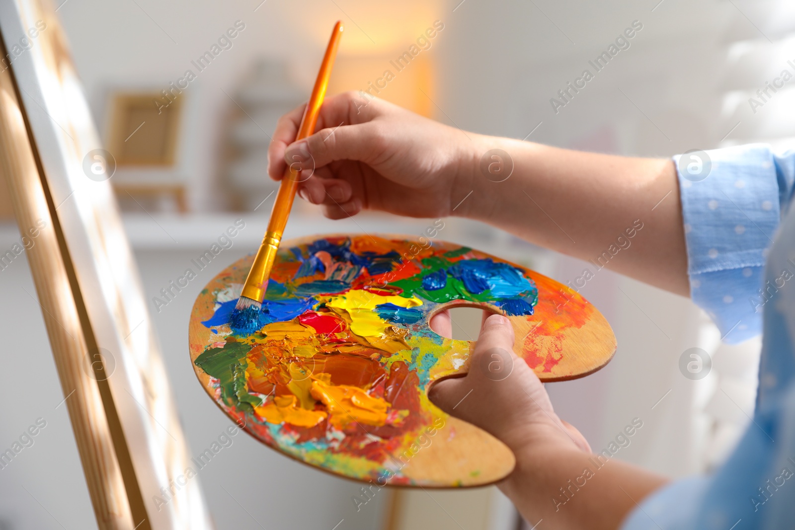 Photo of Woman mixing paints on palette with brush near easel indoors, closeup