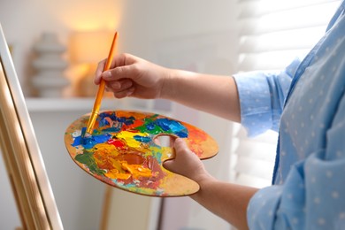 Photo of Woman mixing paints on palette with brush near easel indoors, closeup