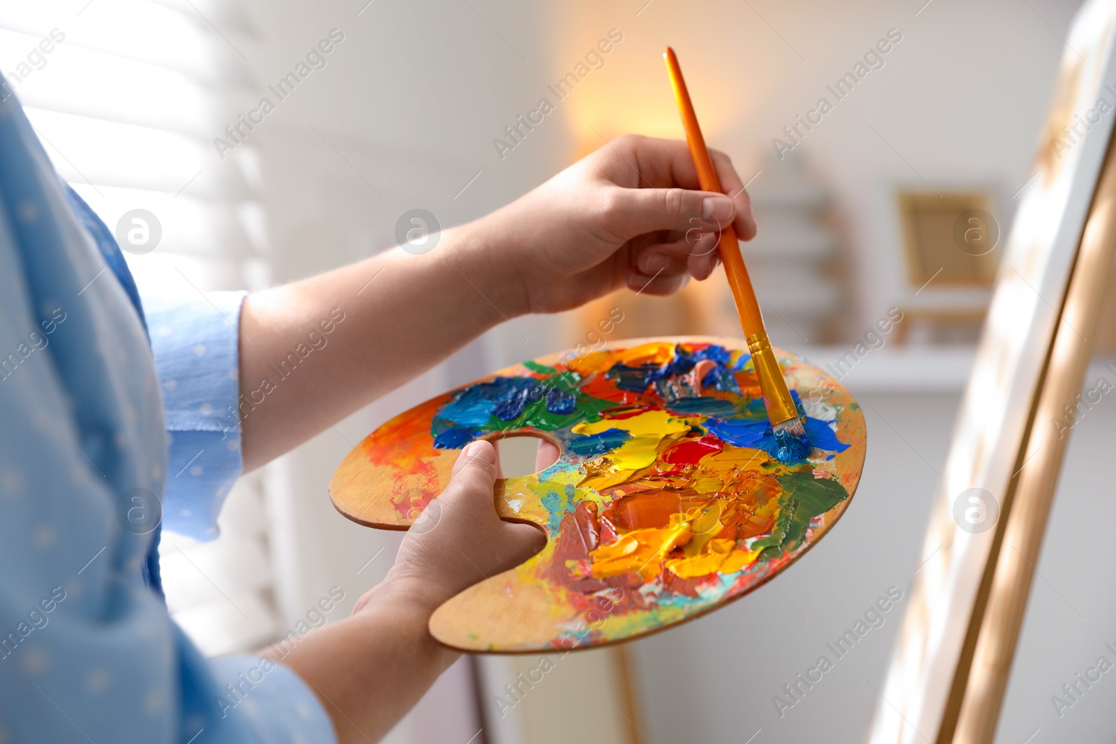 Photo of Woman mixing paints on palette with brush near easel indoors, closeup