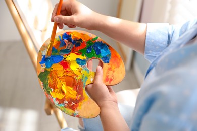 Woman mixing paints on palette with brush near easel indoors, closeup