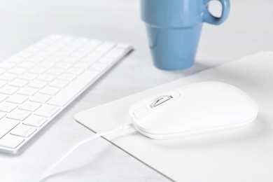 Photo of Computer mouse with mousepad, keyboard and cup on light wooden table, closeup