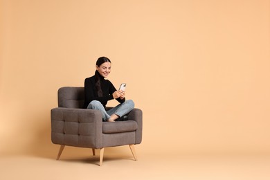 Photo of Smiling woman with smartphone sitting on armchair against beige background. Space for text