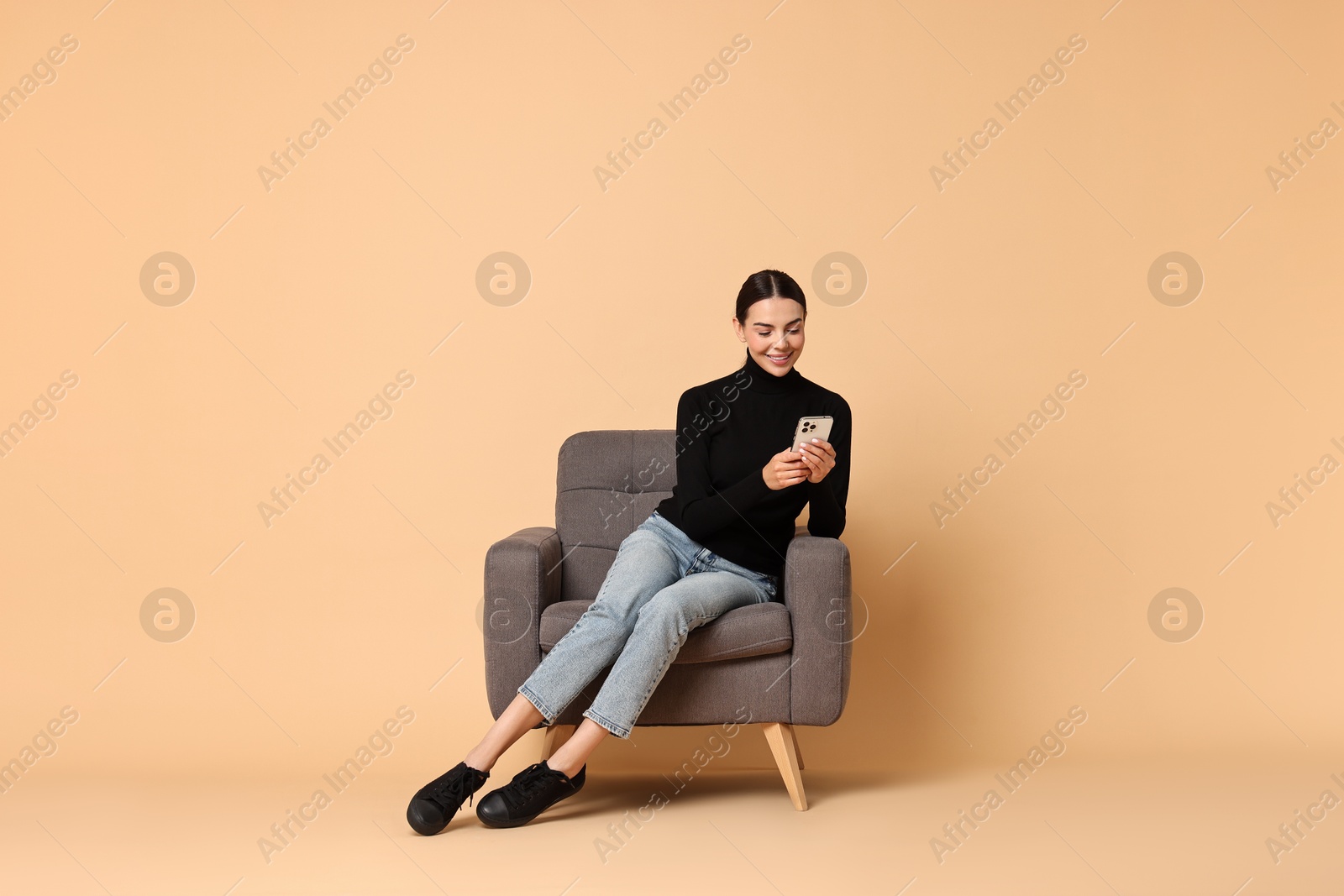 Photo of Smiling woman with smartphone sitting on armchair against beige background