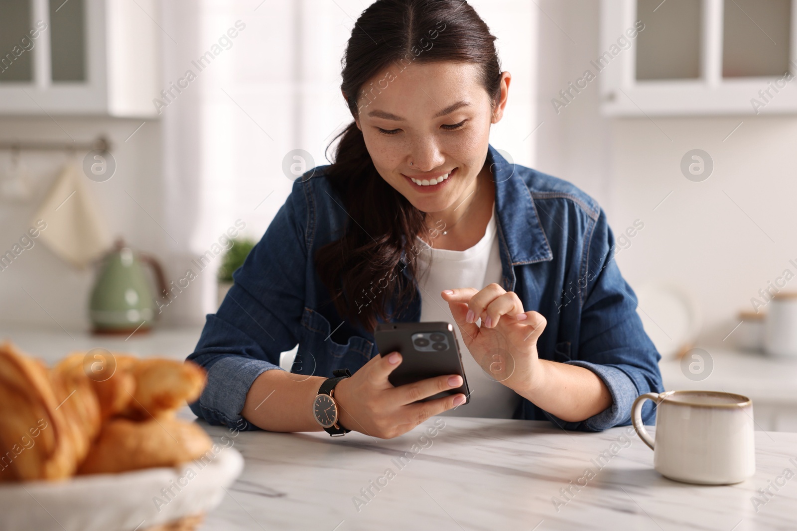 Photo of Smiling woman using smartphone at table in kitchen