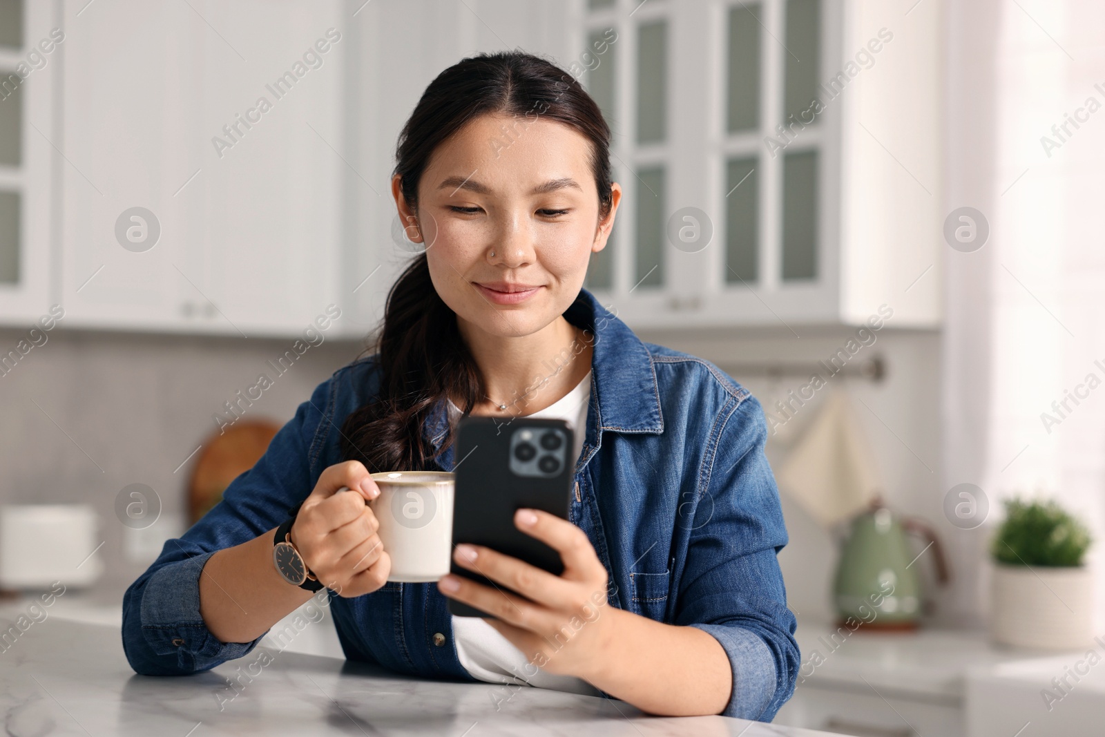 Photo of Beautiful woman with smartphone drinking coffee at table in kitchen