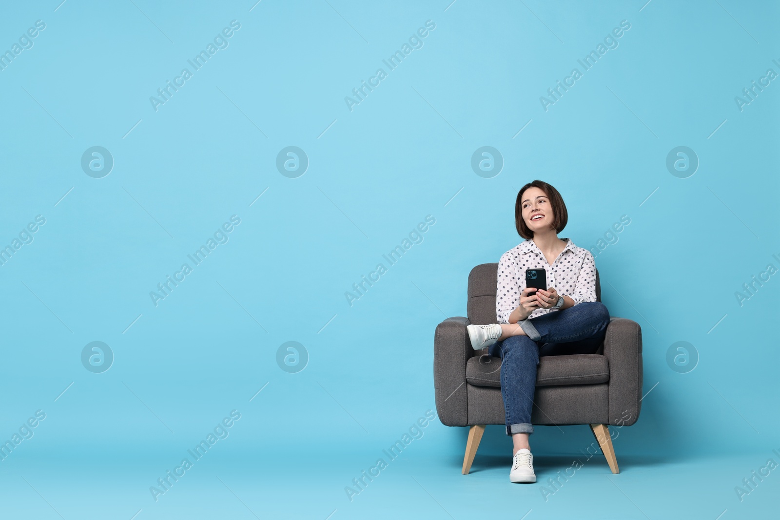 Photo of Smiling woman with smartphone sitting on armchair against light blue background. Space for text