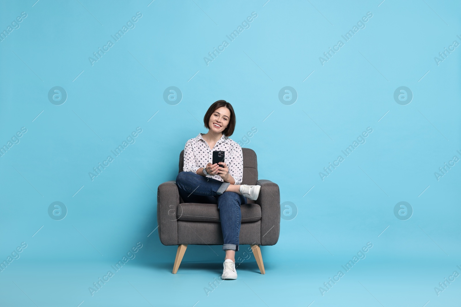 Photo of Smiling woman with smartphone sitting on armchair against light blue background