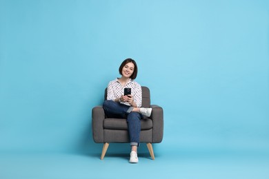 Smiling woman with smartphone sitting on armchair against light blue background