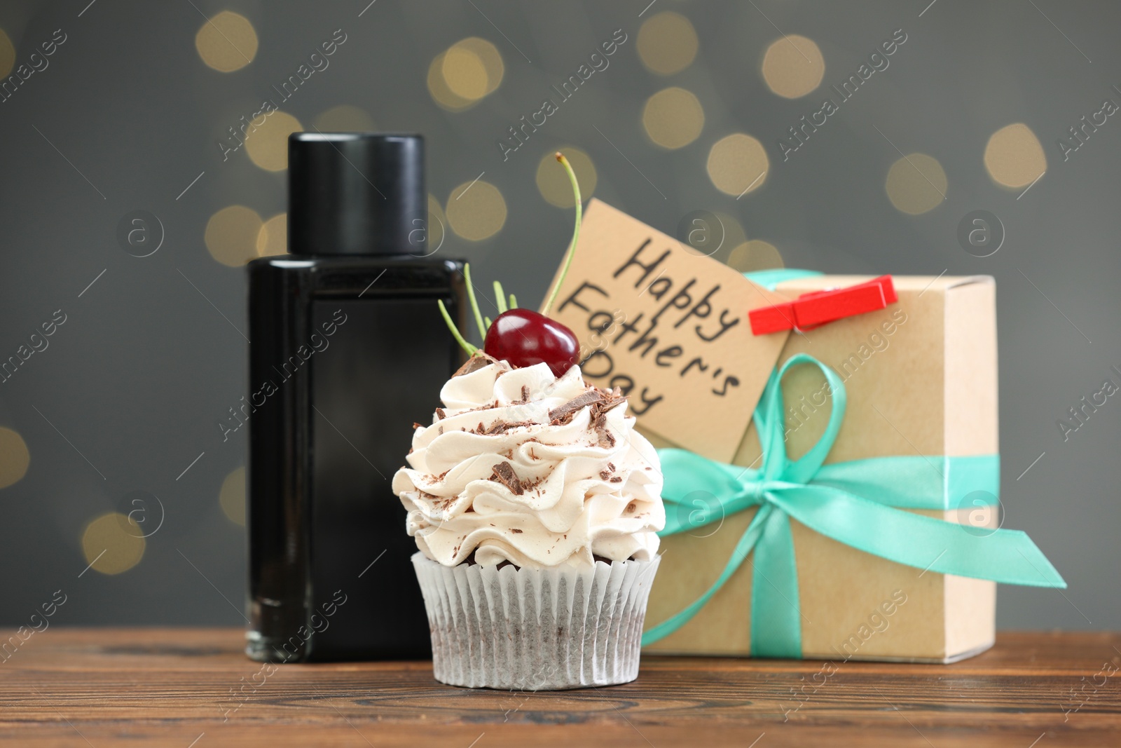 Photo of Greeting card with phrase Happy Father's Day, cupcake, gift box and perfume on wooden table against blurred lights